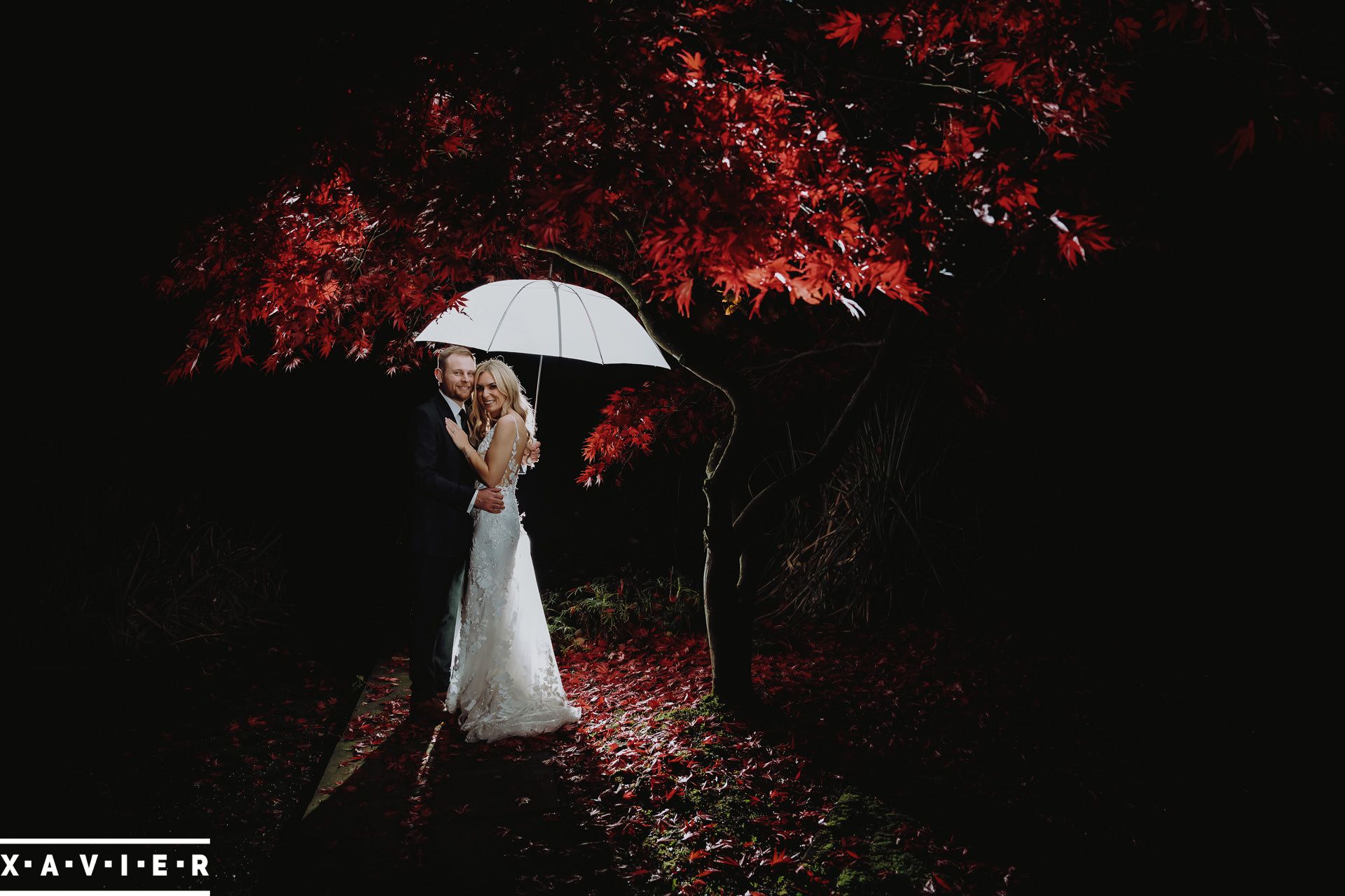 bride and groom stand under white umbrella and red tree in the darl